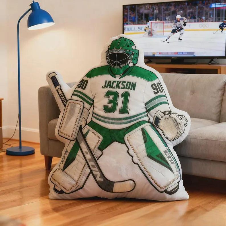 Pode incluir: Uma almofada em forma de guarda-redes de h&oacute;quei, em branco e verde, com o nome "Jackson" e o n&uacute;mero "31". A almofada tem um capacete e prote&ccedil;&otilde;es verdes e est&aacute; colocada num sof&aacute; claro.