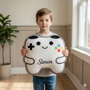 May include: A white, plush video game controller-shaped pillow with black trim and details. The controller features a directional pad, colorful buttons, and the name "Simon" in black script. The pillow is held by a person wearing a striped shirt and jeans.