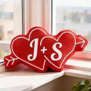 May include: Red heart-shaped cushion with white trim and the letters "J + S" in white. An arrow design is on the left and right sides of the cushion. The cushion is resting on a white windowsill.