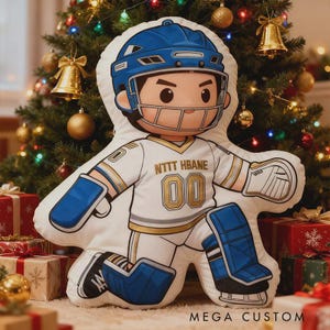 Op de afbeelding: Een kussenvormige hockey speler in een wit en blauw uniform met de tekst "NTTT HBANE 00". Het kussen heeft een blauwe helm, pads en handschoenen. Op de achtergrond een kerstboom en ingepakte cadeaus.