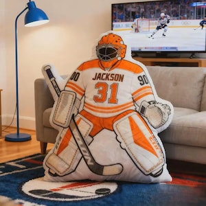 Pode incluir: Uma almofada em forma de guarda-redes de h&oacute;quei em branco e laranja, com o nome "Jackson" e o n&uacute;mero "31" na camisola. A almofada tem um stick e prote&ccedil;&otilde;es de h&oacute;quei, e est&aacute; num sof&aacute;.