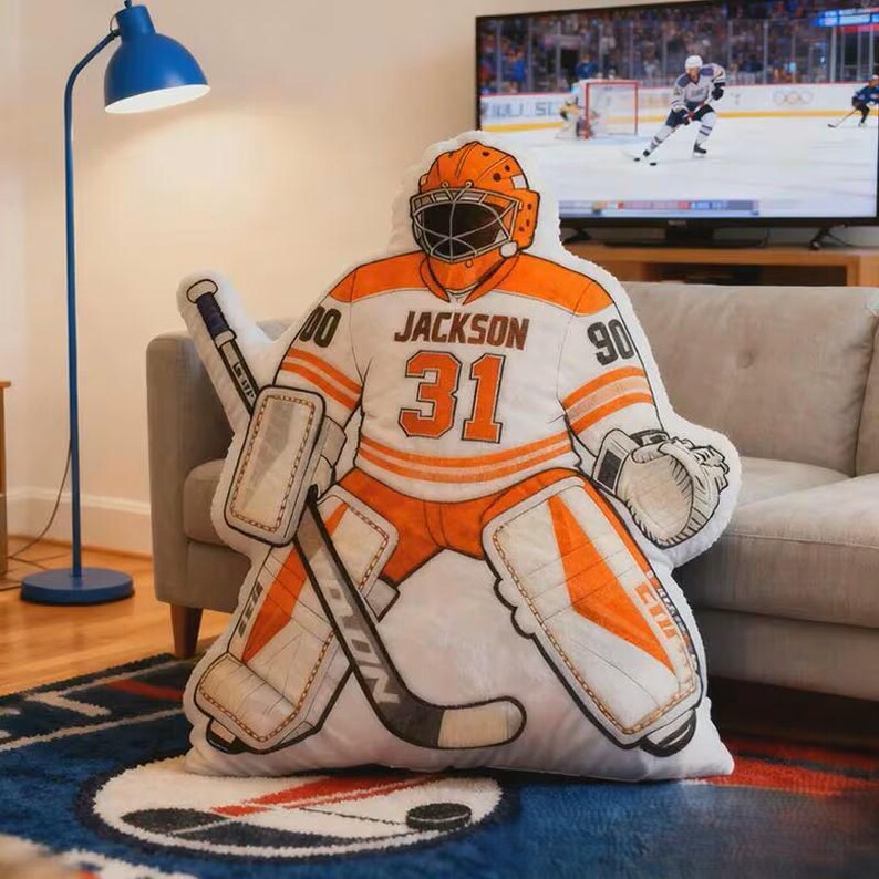 Pode incluir: Uma almofada em forma de guarda-redes de h&oacute;quei em branco e laranja, com o nome "Jackson" e o n&uacute;mero "31" na camisola. A almofada tem um stick e prote&ccedil;&otilde;es de h&oacute;quei, e est&aacute; num sof&aacute;.