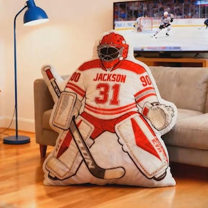 Pode incluir: Uma almofada em forma de guarda-redes de h&oacute;quei em vermelho, branco e cinzento. A almofada apresenta o nome "Jackson" e o n&uacute;mero "31" na camisola. O guarda-redes usa um capacete e prote&ccedil;&otilde;es vermelhas e segura um stick e uma luva de h&oacute;quei.