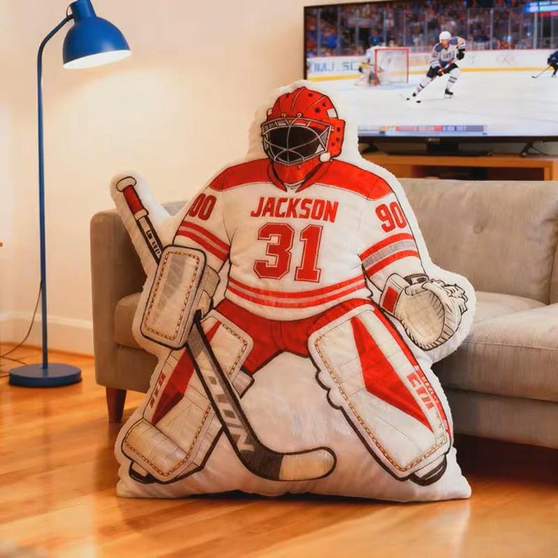 Pode incluir: Uma almofada em forma de guarda-redes de h&oacute;quei em vermelho, branco e cinzento. A almofada apresenta o nome "Jackson" e o n&uacute;mero "31" na camisola. O guarda-redes usa um capacete e prote&ccedil;&otilde;es vermelhas e segura um stick e uma luva de h&oacute;quei.