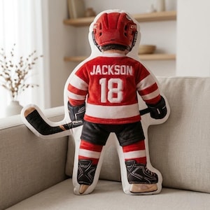 以下が含まれることがあります： 背中に「JACKSON」の名前と「18」の数字が入った赤と白のジャージを着た、ホッケー選手型のクッション。赤いヘルメット、黒いショーツ、ホッケーのスケートも特徴です。ホッケーのスティックが含まれています。