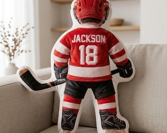 Puede incluir: Un cojín de peluche con forma de jugador de hockey que lleva un casco rojo, una camiseta roja y blanca con el nombre JACKSON y el número 18 impresos en la espalda, pantalones cortos negros y patines negros, sosteniendo un palo de hockey.