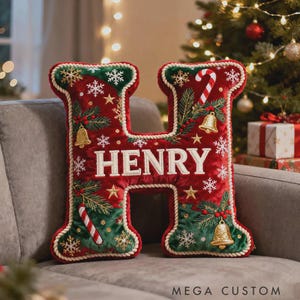 Op de afbeelding: Een decoratief kerstkussen in de vorm van de letter "H". Het rode kussen heeft groene accenten en de naam "HENRY" in het wit geborduurd. Het is versierd met sneeuwvlokken, zuurstokken en gouden bellen.