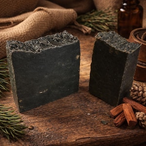 May include: Two dark gray rectangular bars of soap with textured tops are displayed on a wooden surface. The soap is surrounded by pine needles, cinnamon sticks, and a pine cone. A burlap sack and a small brown bottle are in the background.