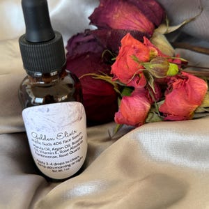 May include: A brown glass bottle with a black dropper, labelled "Golden Elixir Face Serum." The label lists ingredients like Marula Oil and Rosehip. Dried red roses and buds are arranged beside the bottle. Net weight is 28g.