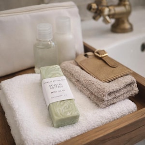 May include: A wooden tray displays bathroom essentials. There's a green soap bar with "Coastal Cottage Mini Soap" text, two small bottles, a white pouch, a tan luggage tag, and folded white and beige towels.