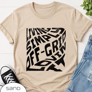 Op de afbeelding: Zandkleurig T-shirt met een zwart grafisch ontwerp. Het ontwerp bevat de woorden "LIVING SIMPLE OFF-GRID NOW" in een vervormd, blokkerig lettertype. Het shirt heeft een ronde hals en korte mouwen. Een zwart-witte sneaker is linksonder zichtbaar.