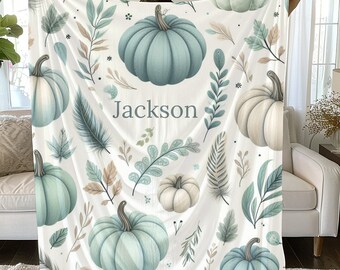 Puede incluir: Una manta de forro polar blanca con el nombre personalizado "Jackson" impreso en el centro. La manta presenta un patrón repetitivo de calabazas azules y blancas, hojas y plumas.
