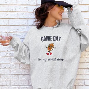 Może przedstawiać: Szara bluza z napisem "GAME DAY is my cheat day" i postacią kreskówkowego piłkarza w czerwonych butach.