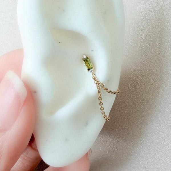 Delicado piercing labret verde musgo em formato de baguete, calibre 16g ou 18g, com pino de pressão, em titânio grau implante hipoalergênico. Também pode ser usado com argola, piercing labret, piercing de concha ou corrente para concha. Base plana.