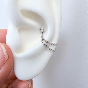 Puede incluir: Un pendiente tipo ear cuff de tono plateado con un dije de estrella de cuatro puntas y una pequeña piedra iridiscente en el centro. Dos cadenas plateadas finas cuelgan de la estrella y se conectan a otro punto del pendiente.
