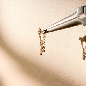 May include: A gold earring with a small blue gemstone and dangling chains with tiny gold beads is held by a silver tool. The earring is suspended against a beige background with shadows.