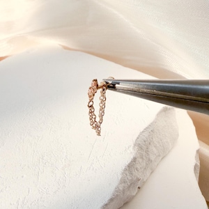 Puede incluir: Un delicado pendiente de cadena dorada con un pequeño perno y una cadena colgante. El pendiente está sujeto por unos alicates contra una superficie blanca texturizada y un fondo de tela color crema.