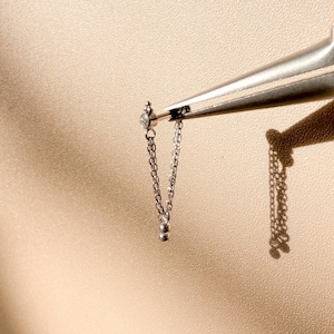 Könnte beinhalten: Ein kleiner, silberner Ohrring mit einer hängenden Kette und einem kleinen, klaren Edelstein. Der Ohrring wird von einer Zange vor einem neutralen Hintergrund gehalten, was sein filigranes Design und seine Handwerkskunst zeigt. Die Kette hat einen kleinen, runden Anhänger am Ende.