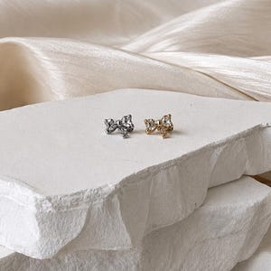 May include: Two small stud earrings shaped like bows, one in a silver tone and one in a gold tone, each featuring a clear, faceted stone at the center. The earrings are positioned side by side on a textured, white, rectangular stone surface.