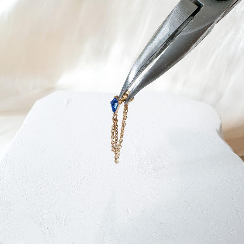 May include: A gold earring with a small, blue gemstone and a dangling chain is held by pliers. The earring is set against a white background, highlighting the jewelry's delicate design. The earring is a stylish accessory. The chain is approximately 2.5 cm long.