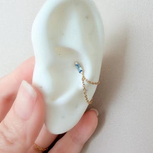May include: A gold-colored ear cuff featuring a rectangular, light blue gemstone and a delicate chain. The cuff is designed to wrap around the ear, creating a stylish and modern look. The jewelry is shown on a white ear model.