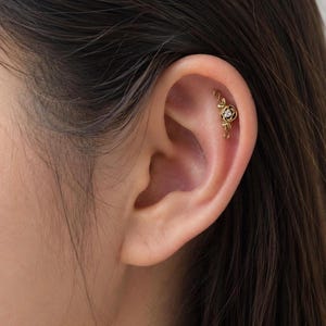 May include: A gold-colored helix earring featuring a floral design with a small, clear gemstone at the center. The earring is shaped like a rose with delicate leaves and is worn on the upper ear cartilage.