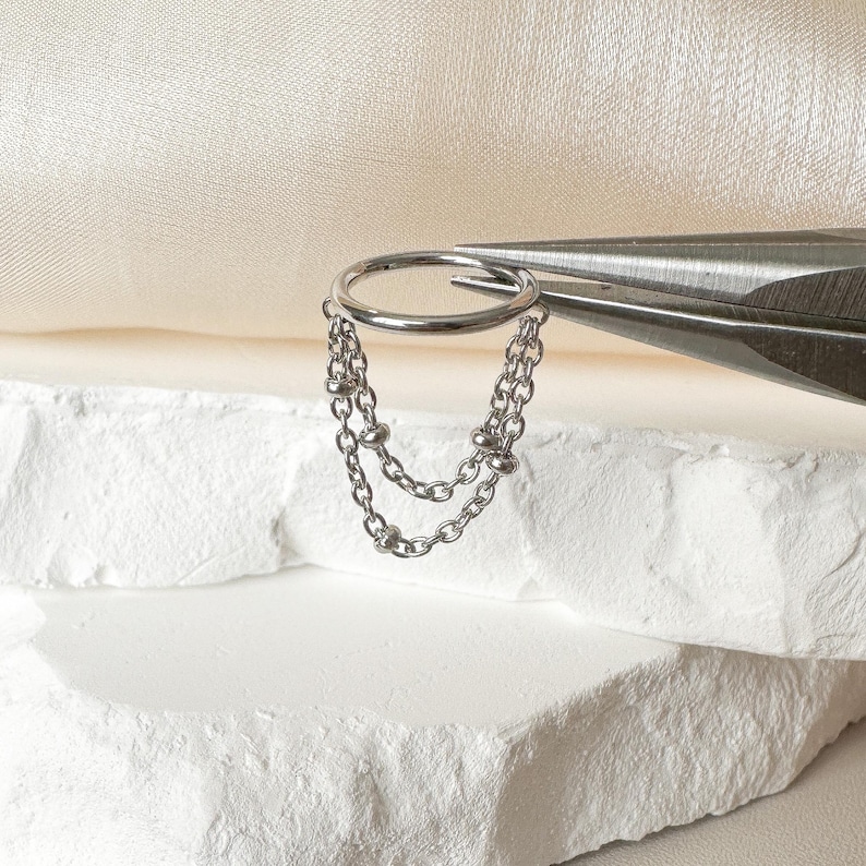 May include: A silver-colored hoop earring with two dangling chain strands. The earring is held by a pair of silver metal pliers. The earring is displayed on a white, textured surface.