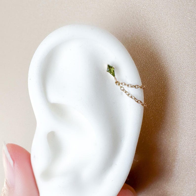 May include: A gold ear cuff with a small, green, diamond-shaped gemstone linked by a fine chain to another point on the ear. The jewellery is displayed on a white ear model.