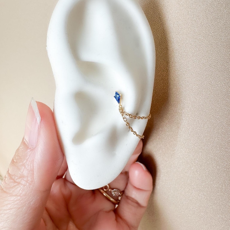 May include: A gold ear cuff with a small, blue, diamond-shaped gemstone. A delicate gold chain connects the gemstone to the ear cuff. The jewelry is displayed on a white ear model, with a hand in the foreground.