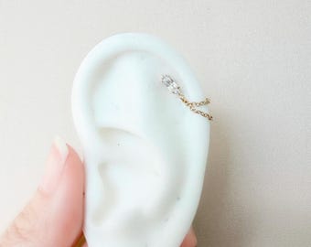 May include: A gold ear cuff with a small, clear, rectangular stone and a fine chain. The chain hangs from the stone to the ear's edge. The jewellery is shown on a white ear model.