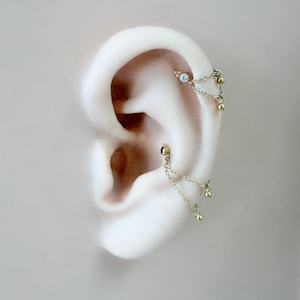 May include: Gold-toned ear piercings with dangling chain and bead accents. One piercing features a small, clear gemstone. The jewelry is displayed on a light-colored ear model, showcasing the design and details.