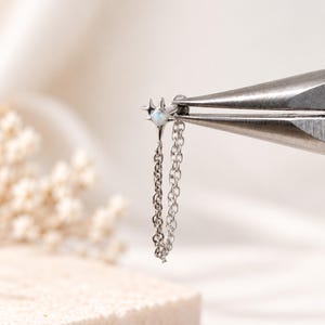 May include: A silver-toned body jewelry piece featuring a star-shaped charm with a small, iridescent white stone at its center. Two delicate, thin silver chains hang from the star charm, creating a draped effect for this piece of jewelry.