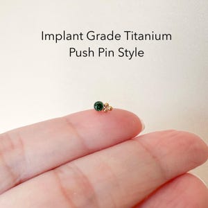 Puede incluir: Un pendiente de estilo pinza de titanio con una piedra preciosa verde y detalles dorados. Titanio de Grado Implantable Estilo Pinza.