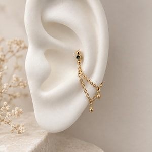 May include: A gold-toned ear cuff featuring a small dark green gemstone set in a metal bead, connected by two delicate link chains to a dangling gold bead. The jewelry piece is positioned on the upper cartilage of a human ear structure.