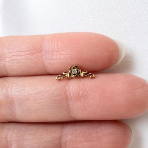 Può includere: Un piccolo gioiello color oro con un design a rosa e un diamante centrale. La rosa è affiancata da delicati accenti di foglie e viti curve. Il gioiello è mostrato su uno sfondo neutro.