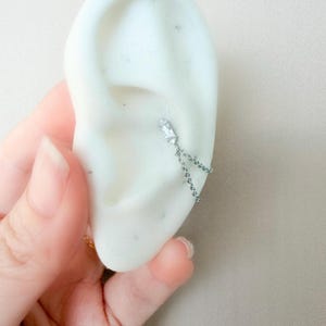 Puede incluir: Un pendiente plateado con una gema rectangular y transparente y una delicada cadena. El pendiente está sujeto a un modelo de oreja blanco, con una mano sosteniendo el modelo en el fondo.