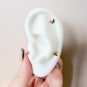 May include: Gold-colored butterfly stud earrings displayed on a white ear model. The earrings are small and delicate, with three visible on the ear. The ear is held by a hand with a black and silver nail design.