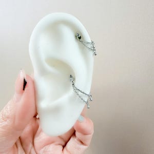 May include: Silver chain earrings with dangling beads and a small gem. The earrings are shown on a white ear model. The earrings are designed to be worn in multiple ear piercings.