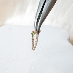 May include: A gold-coloured earring with a green gemstone and a dangling chain is held by a pair of pliers. The earring features a small, square-cut green stone and a delicate chain that hangs down.