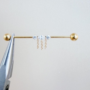 May include: A gold industrial barbell earring with three clear, sparkling gems and dangling chains. The barbell has a ball on each end. The earring is held by a silver tool against a white background.