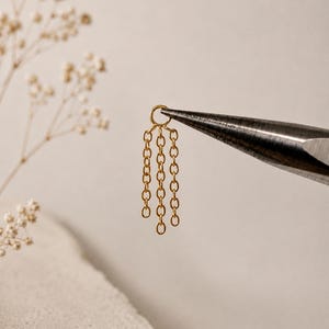 May include: A gold-toned metal jewelry component featuring a small circular jump ring attached to three dangling chain segments of varying lengths. The piece is held by a pair of metal pliers, showcasing the delicate link structure and metallic finish.