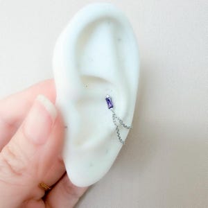 May include: A silver ear piercing featuring a rectangular, purple gemstone and a delicate chain. The jewelry is displayed on a white ear model, showcasing the design and style of the earring. The chain dangles gracefully.
