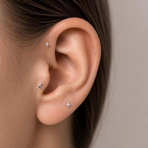 Könnte beinhalten: Nahaufnahme eines Ohrs mit drei kleinen, silbernen Ohrringen. Zwei sind sternförmig, einer ist ein Halbmond. Die Ohrringe befinden sich in Helix, Tragus und Ohrläppchen.