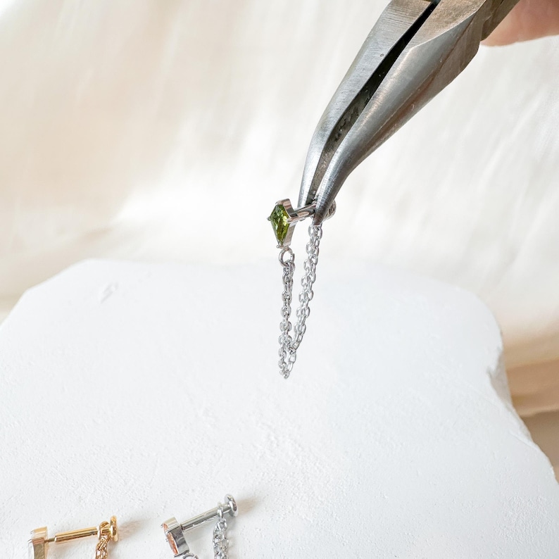 May include: A silver earring with a green gemstone and a dangling chain is held by pliers. The earring is set against a white background, with two other earrings visible at the bottom.