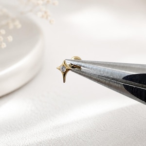 May include: A gold-toned stud earring featuring a four-pointed star shape with a small, clear, round-cut gemstone set in the center. The earring is held by a pair of metal pliers, showing the post and flat back component of the jewelry piece.
