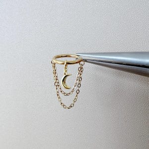 Op de afbeelding: Een goudkleurige ring met een maansikkelbedel en twee hangende kettingen. De ring wordt vastgehouden door een zilverkleurige pincet tegen een neutrale achtergrond. Een delicaat sieraad.