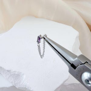 May include: A silver earring featuring a rectangular, purple gemstone and a fine chain. The earring is presented on a white, textured surface. Pliers are visible in the foreground.
