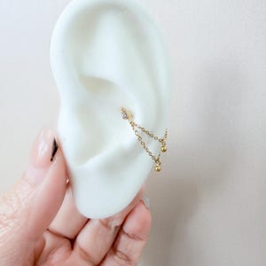 Puede incluir: Un piercing de oreja dorado con un pequeño diamante y una cadena con dos esferas doradas colgantes. La joya se exhibe en un modelo de oreja blanco, mostrando el diseño y los detalles del pendiente.