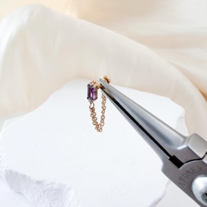 May include: A gold earring featuring a rectangular, purple gemstone and a small diamond. A fine gold chain hangs from the earring. The earring is held by pliers.