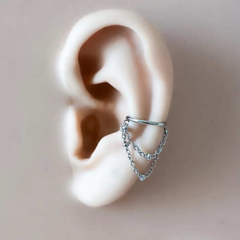 May include: A silver ear cuff featuring a chain design with three dangling strands. The cuff is attached to a model ear, showcasing the jewelry's placement. The ear is set against a neutral background, highlighting the cuff's intricate details and metallic shine.